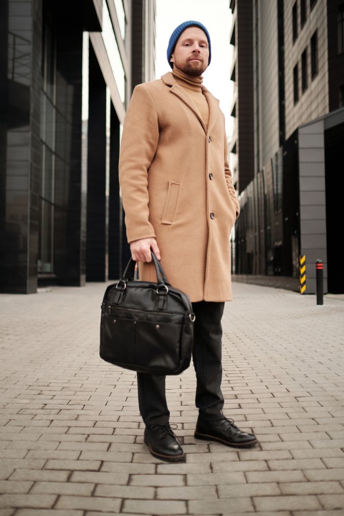 Young businessman in a brown coat standing with a black leather bag in an urban setting.