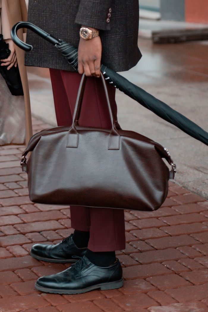 Mastering the First Impression: Your intriguing post title goes here Stylish man holding an umbrella and a leather bag on a paved walkway, showcasing urban fashion.