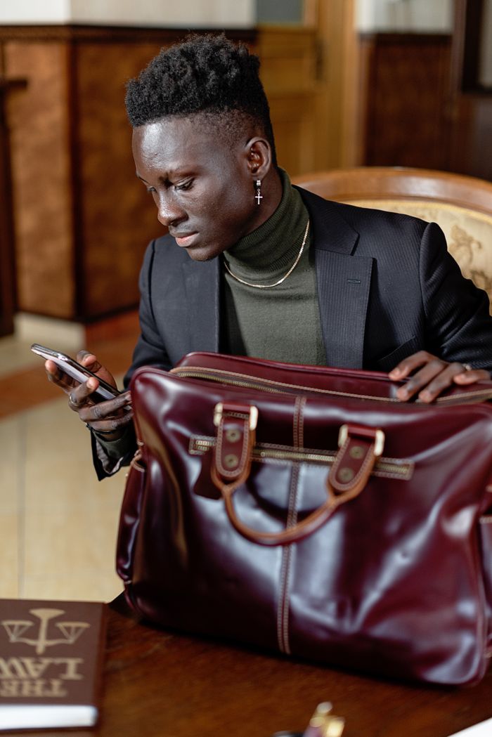 Young professional man in stylish attire using a smartphone while handling a leather bag.